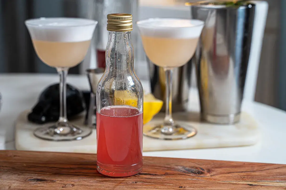 En flaska med rosa rabarbersirap står framför två drinkar med skum. En shaker och en citronklyfta syns i bakgrunden.