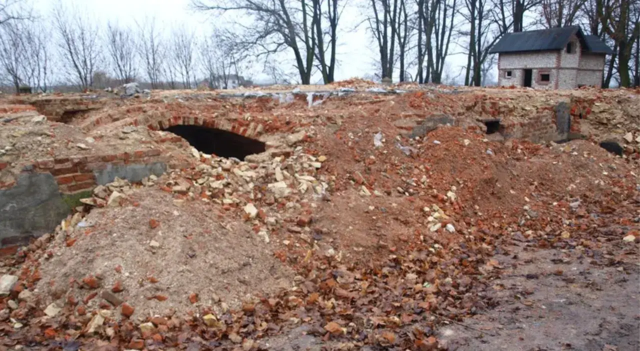 Ruiny ceglane z łukiem i mały domek w tle, otoczone drzewami.