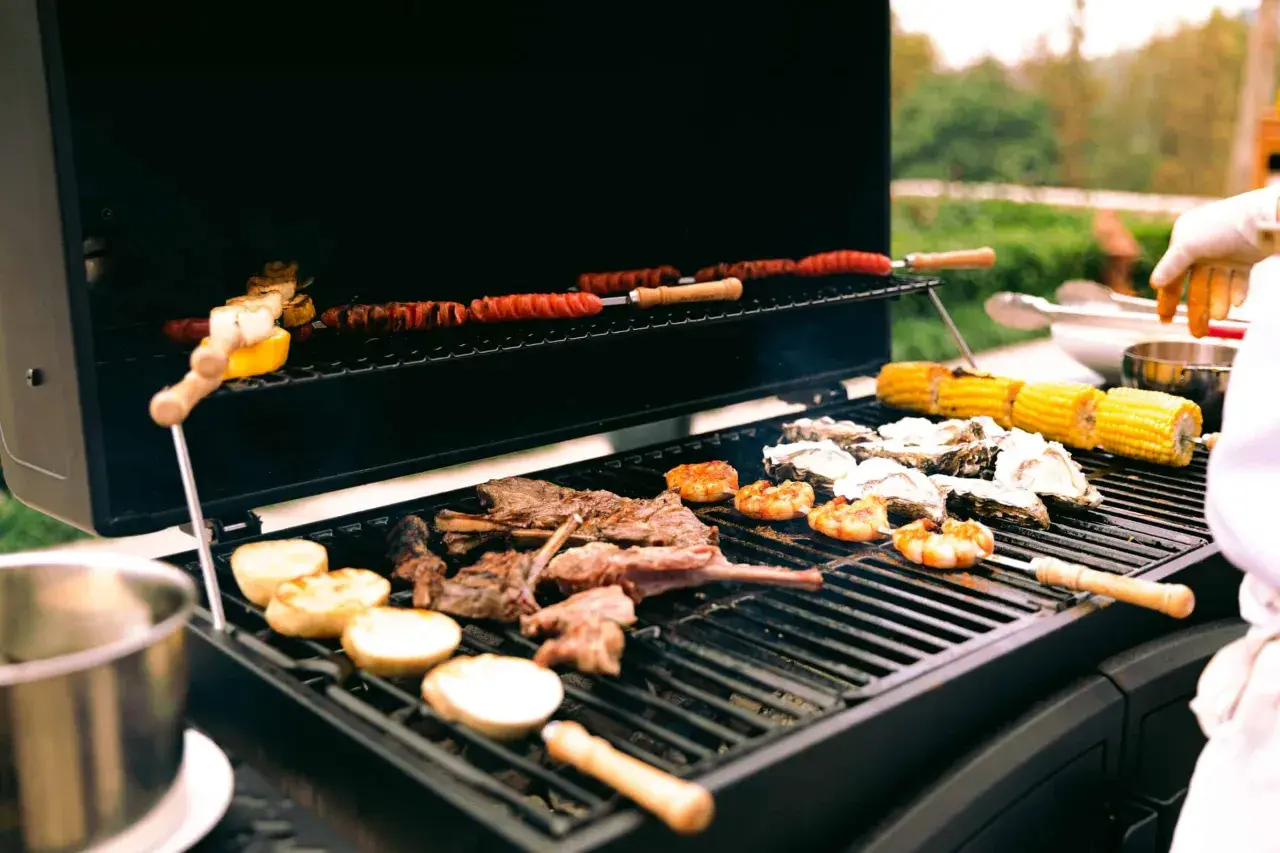 Viande, crevettes, huîtres et maïs grillés sur un barbecue gaz ou charbon. Des saucisses enroulées cuisent sur la grille supérieure.
