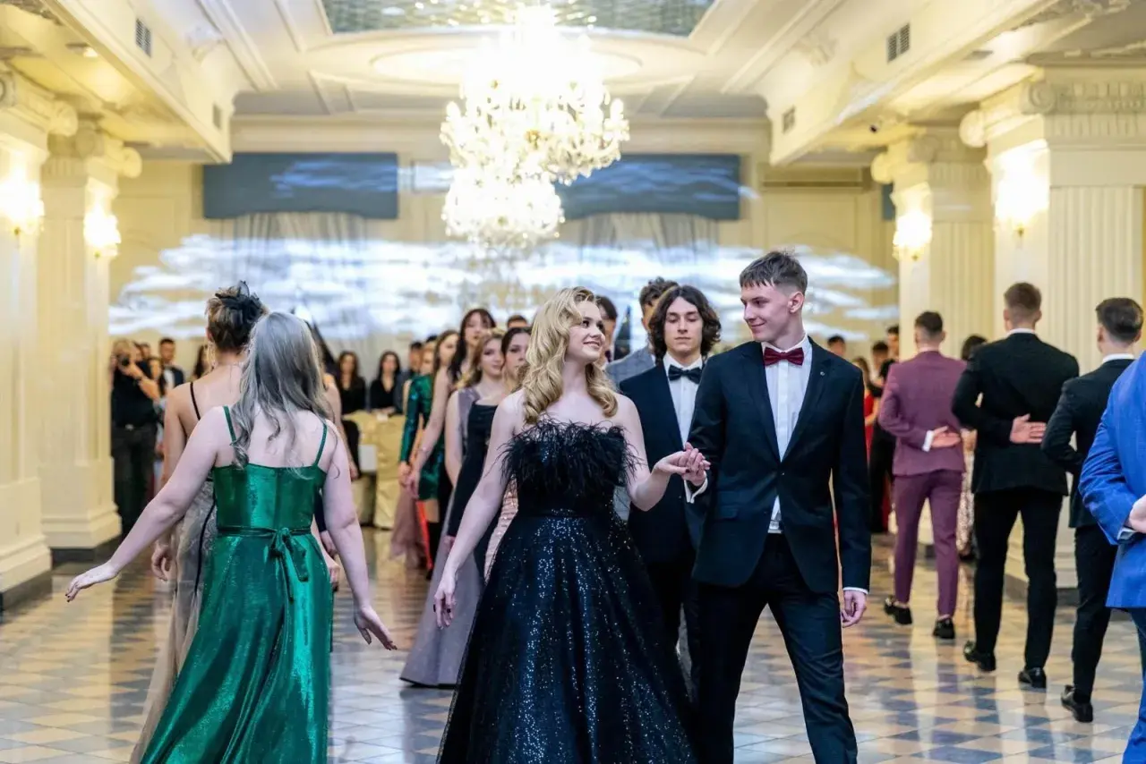 Młoda para idzie na studniówkę. Dziewczyna w czarnej, błyszczącej sukni z piórami, chłopak w eleganckim garniturze. W tle inni uczniowie w balowych strojach.