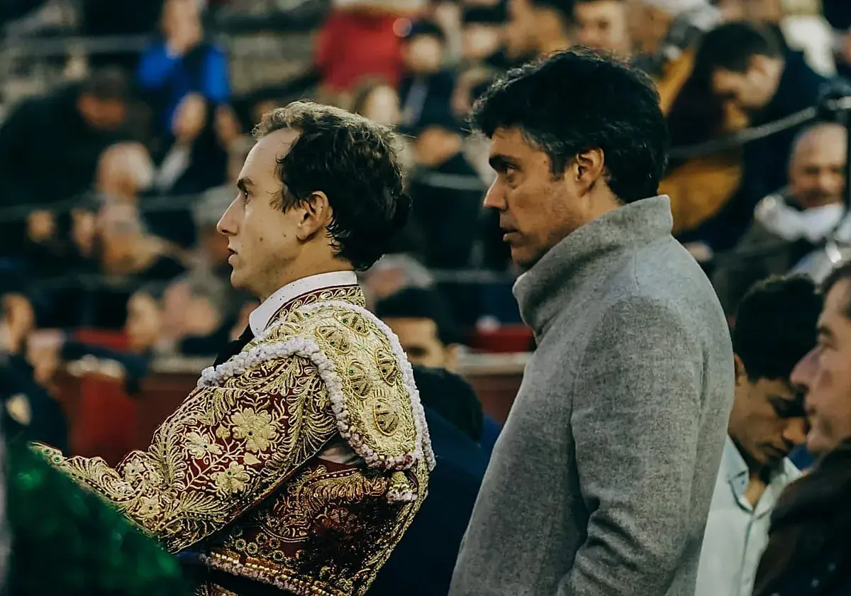 Un torero con traje de luces bordado y zapatillas torero, junto a un hombre con abrigo gris, observan la plaza.