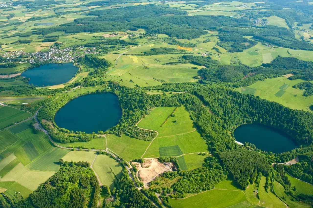 Luftaufnahme des **rundwegs um die drei Dauner Maare**. Grüne Felder, Wälder und drei dunkle Maare prägen die Landschaft.