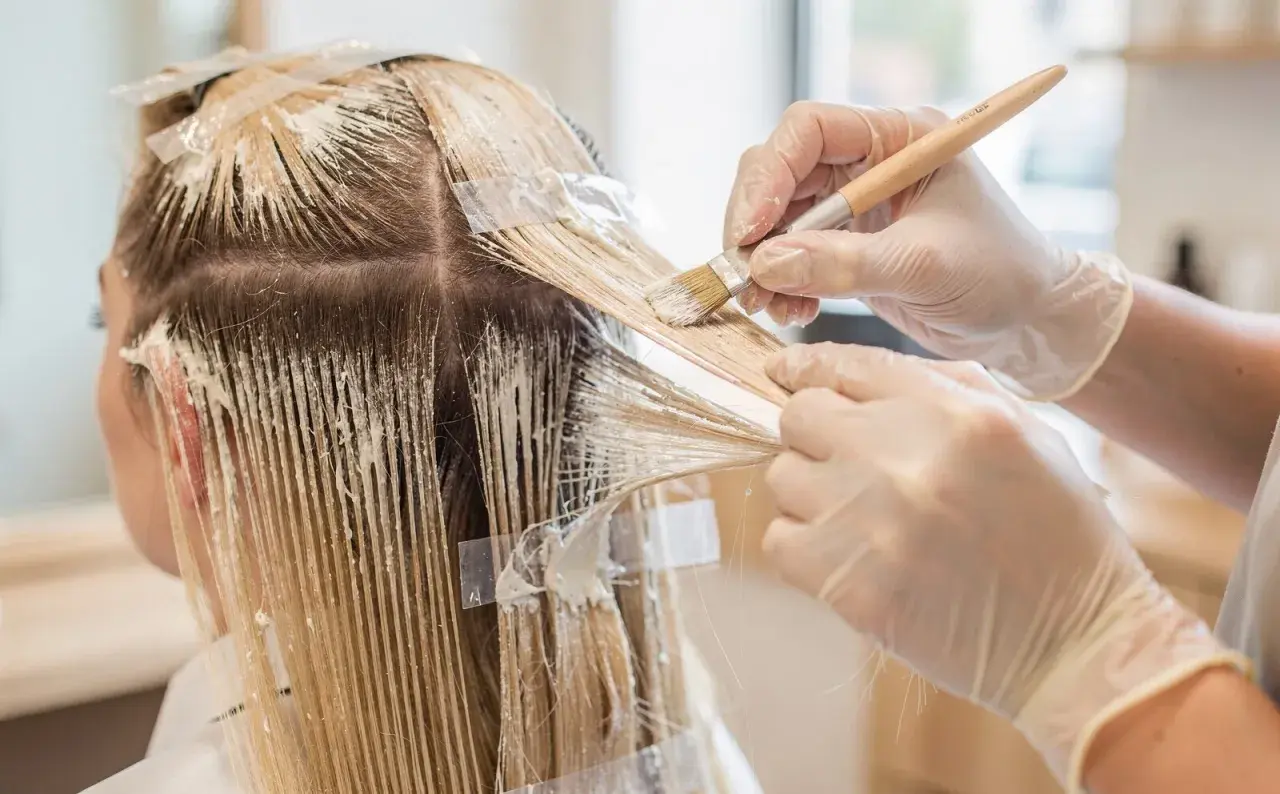 Manos enguantadas aplican tinte para crear mechas blancas en cabello rubio.