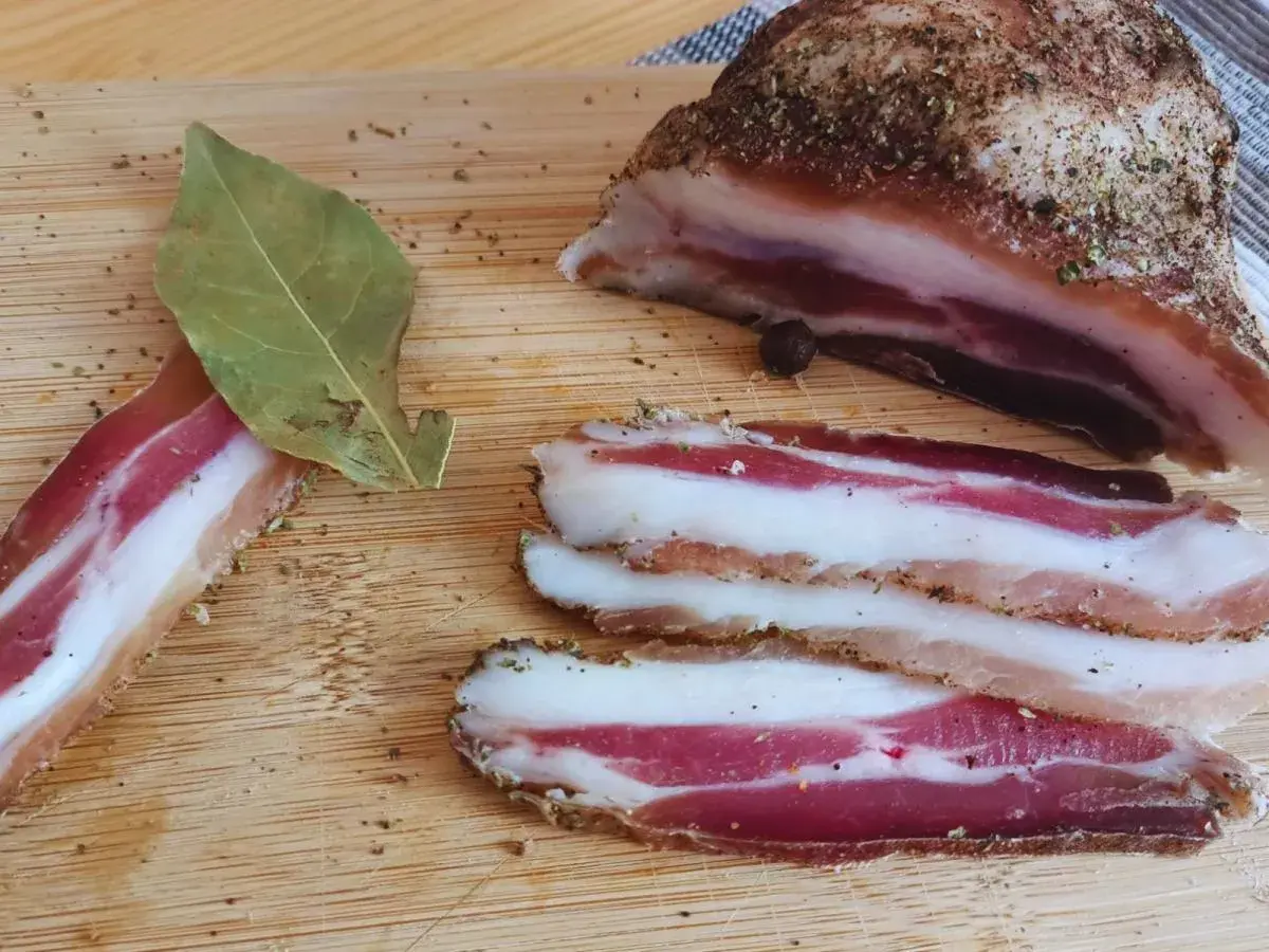 Pancetta co to? Pokrojony w plastry, przyprawiony boczek na desce. Obok liść laurowy i kawałek większego kawałka.