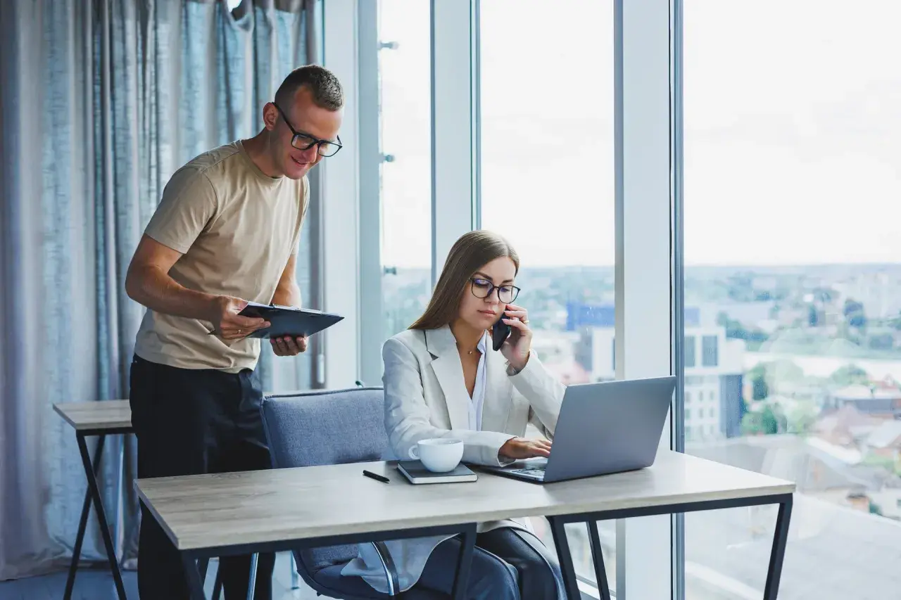 Un homme consulte un document pendant qu'une femme au téléphone travaille sur son ordinateur portable, illustrant l'efficacité de l'e-commerce B2B.