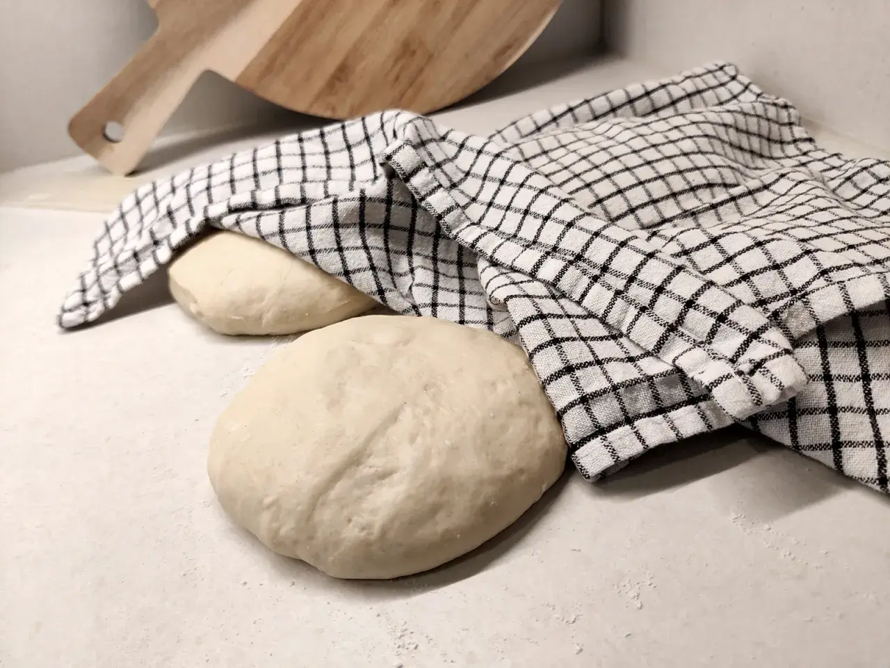 Två runda pizzadegar ligger på en bänk, en täckt av en rutig kökshandduk. Redo för bakning samma dag.