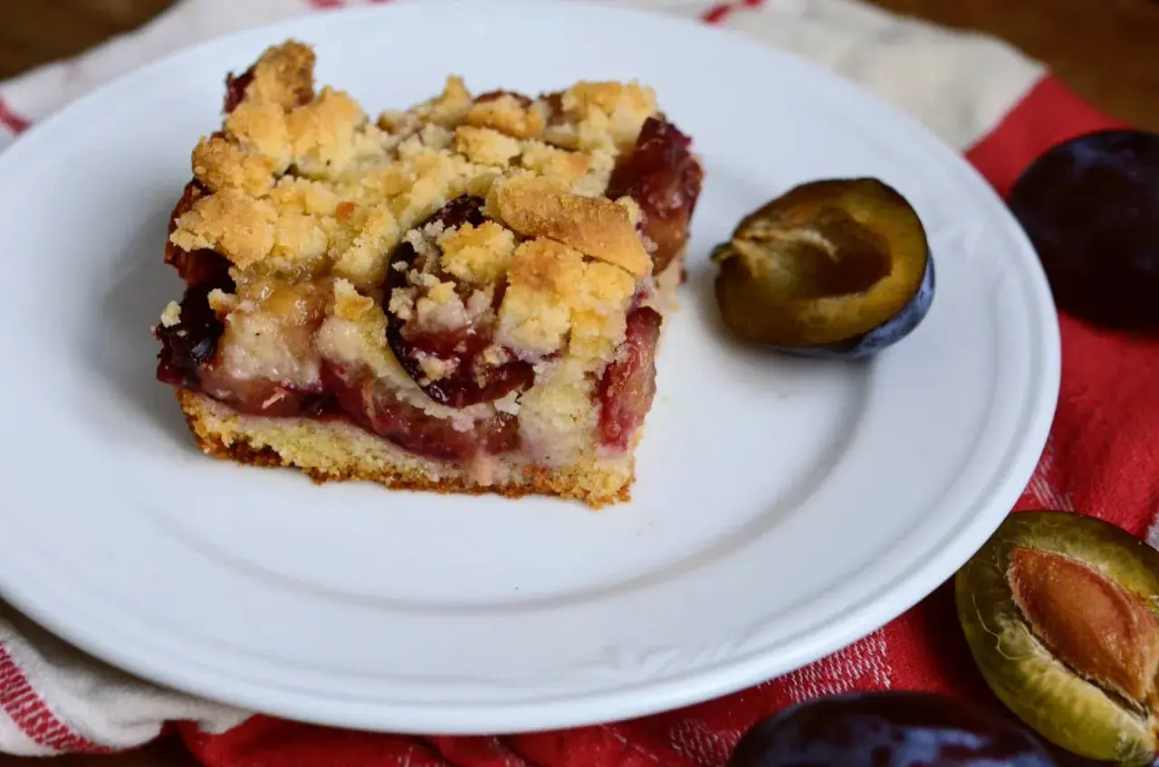 Tranche de gâteau aux prunes à la poêle, garnie de crumble doré et de fruits juteux. Une demi-prune à côté.