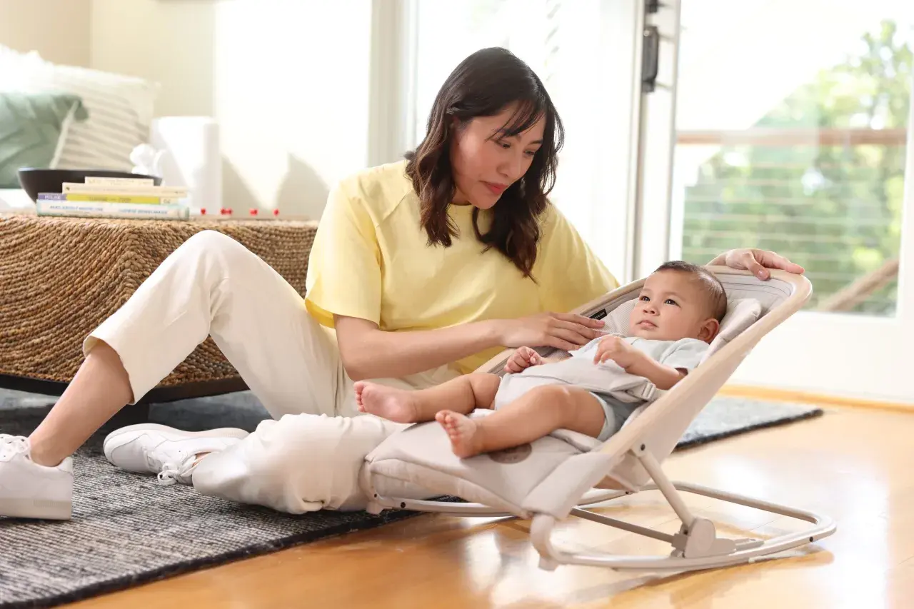 En kvinna sitter på golvet bredvid en bebis i en babysitter från Maxi-Cosi.