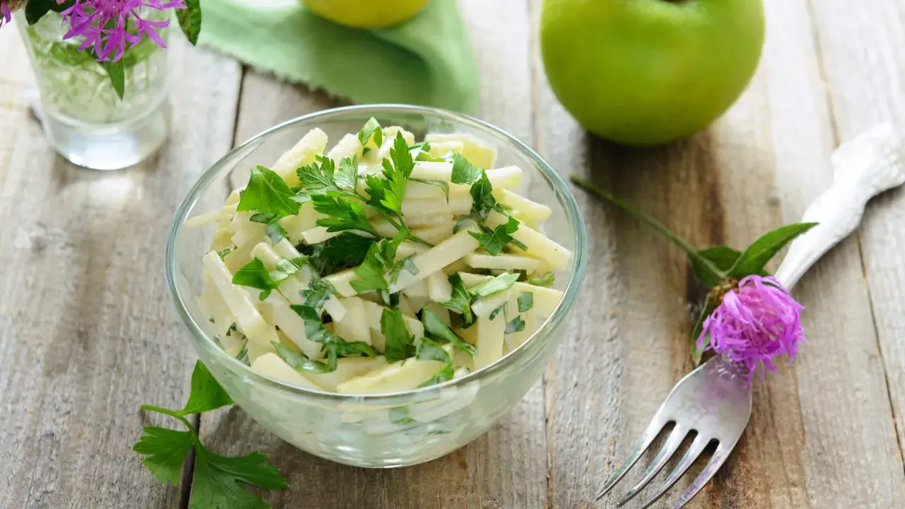 Frischer Kohlrabi Salat mit Petersilie in einer Glasschüssel, garniert mit einer lila Blume und grünen Äpfeln im Hintergrund.