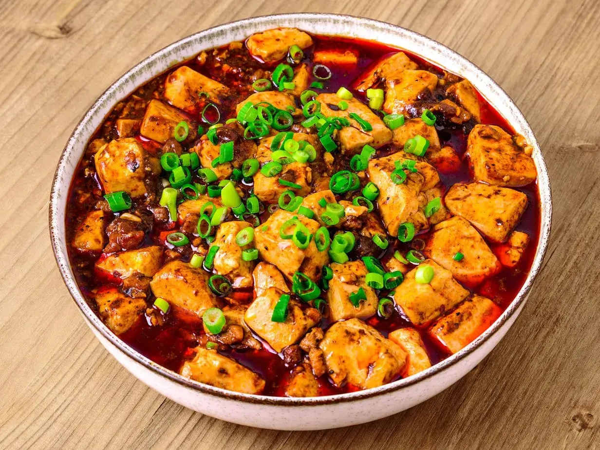 En skål med kryddig mapo tofu, toppad med färsk salladslök, serverad på ett träbord.