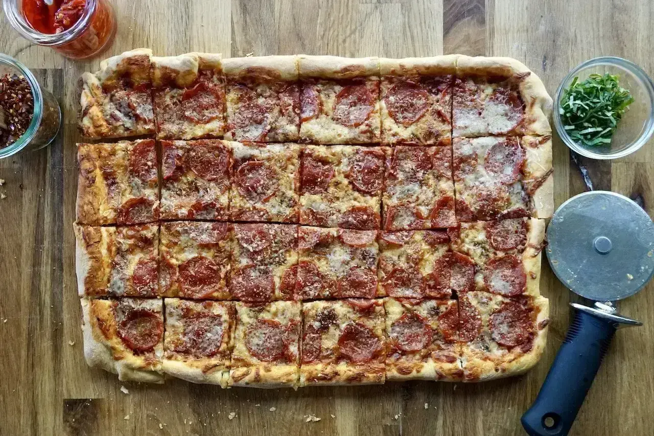 Prosty przepis na pizzę: prostokątna pizza pepperoni z serem, pokrojona na kwadraty, obok słoiczki z dodatkami i krajalnica.
