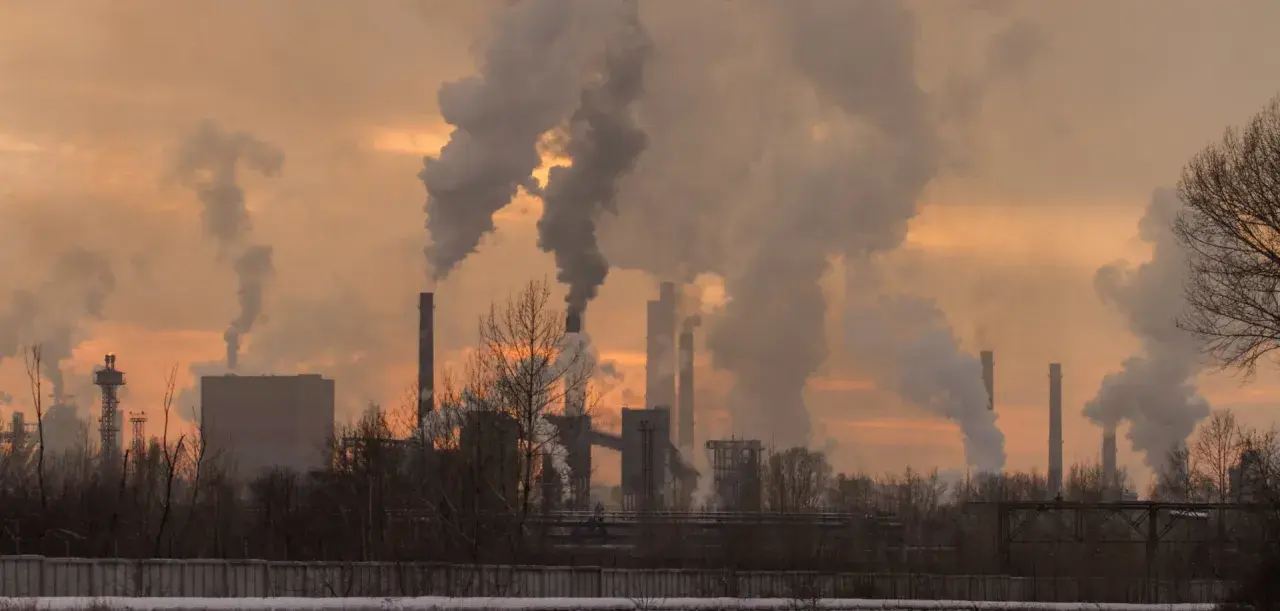 Chimeneas industriales expulsan humo denso, creando un paisaje sombrío. Las partículas contaminantes se elevan, tiñendo el cielo de un naranja opaco.