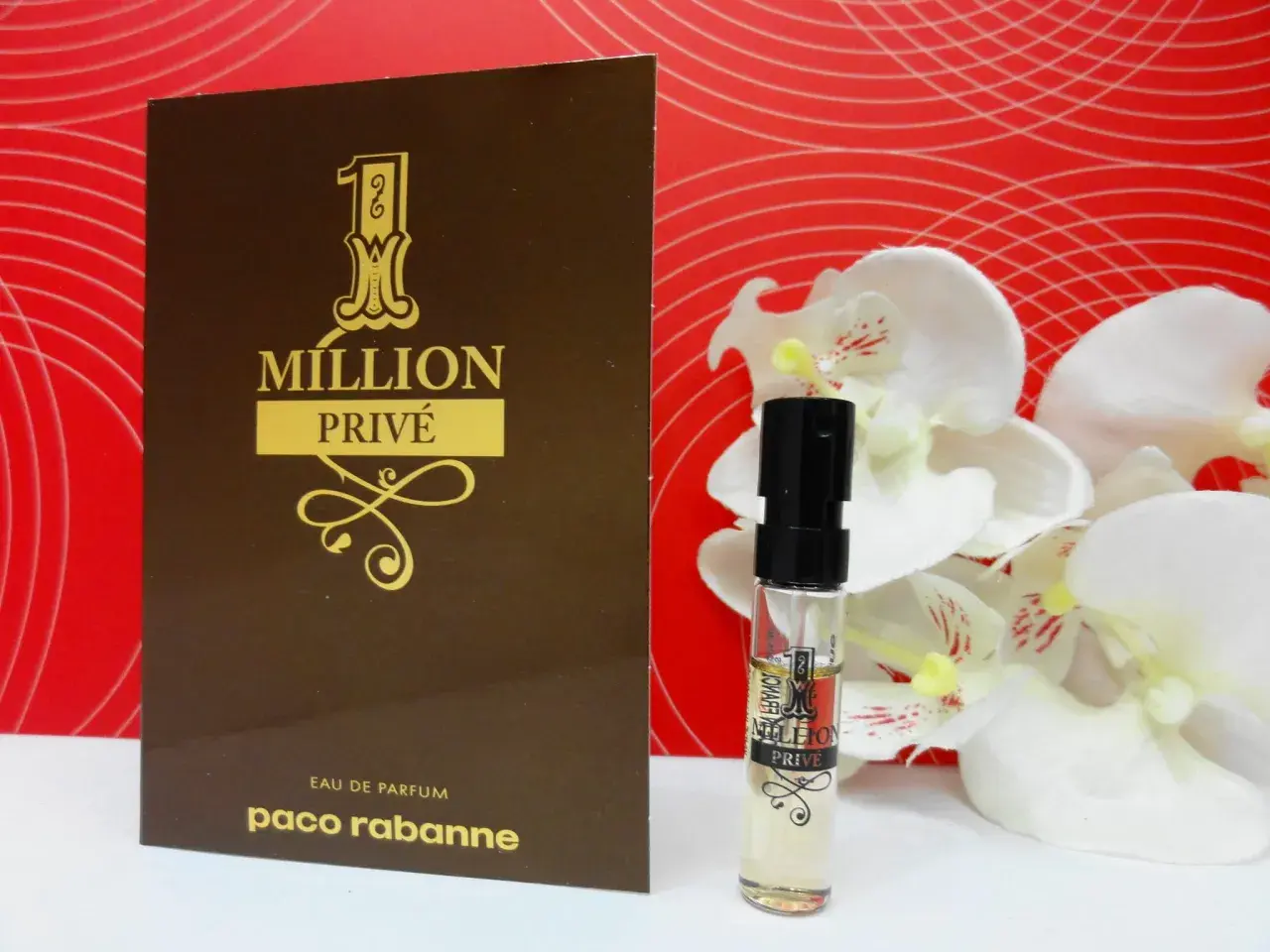 Złoty flakonik i próbka Paco Rabanne 1 Million Privé na tle czerwonych fal i białych kwiatów.
