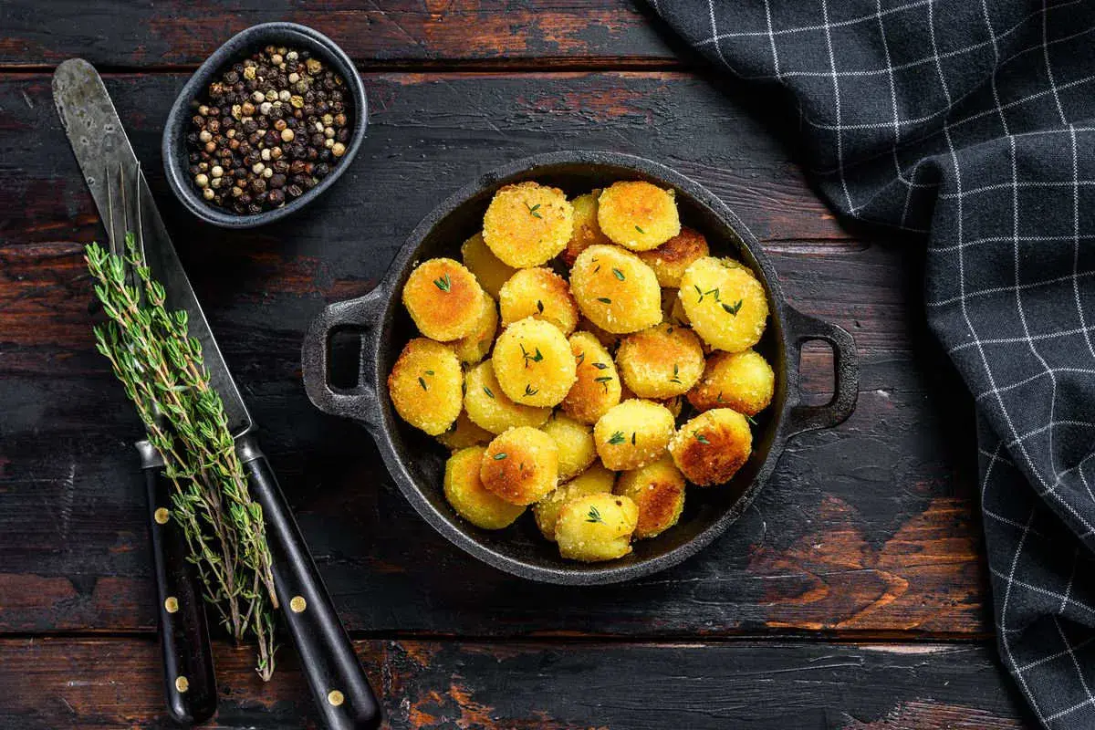 Crispy golden gnocchi in a black pan, garnished with herbs, on a wooden table with peppercorns and a knife.
