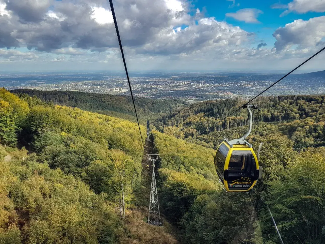 Żółta kolejka linowa z Szyndzielni na Klimczok mija jesienne lasy, a w oddali widać panoramę miasta.