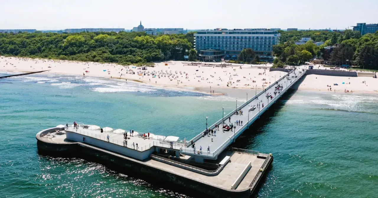 Molo i plaża w słoneczny dzień. W tle widać budynek sanatorium nad morzem, otoczony zielenią.