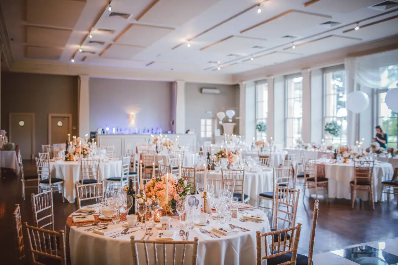 Elegancka sala imprez okolicznościowych przygotowana na przyjęcie. Okrągłe stoły nakryte białymi obrusami, ozdobione kwiatami i świecami.