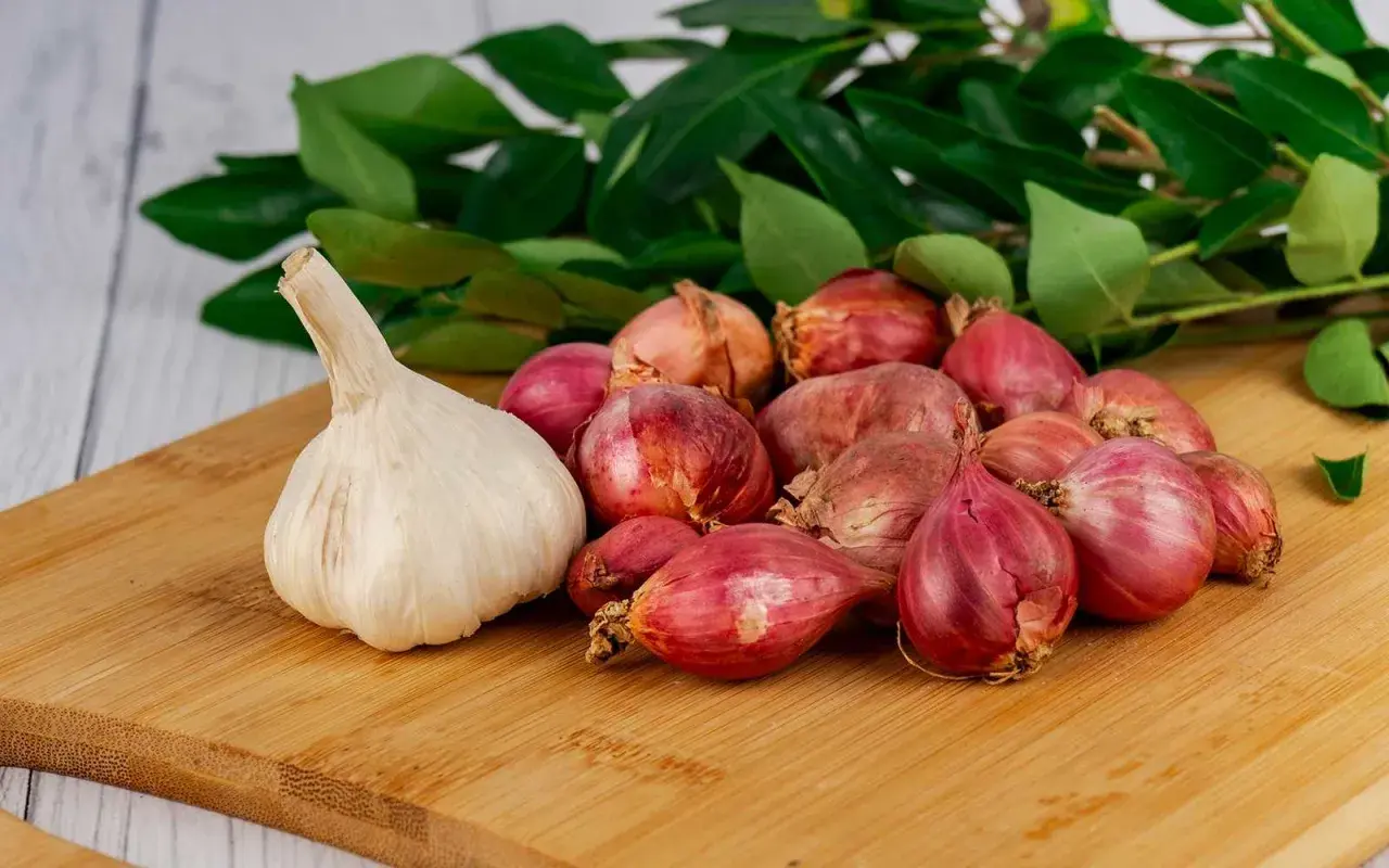 Knoblauch und Schalotten auf einem Holzbrett, bereit als Hausmittel gegen Ohrenschmerzen. Grüne Blätter im Hintergrund.