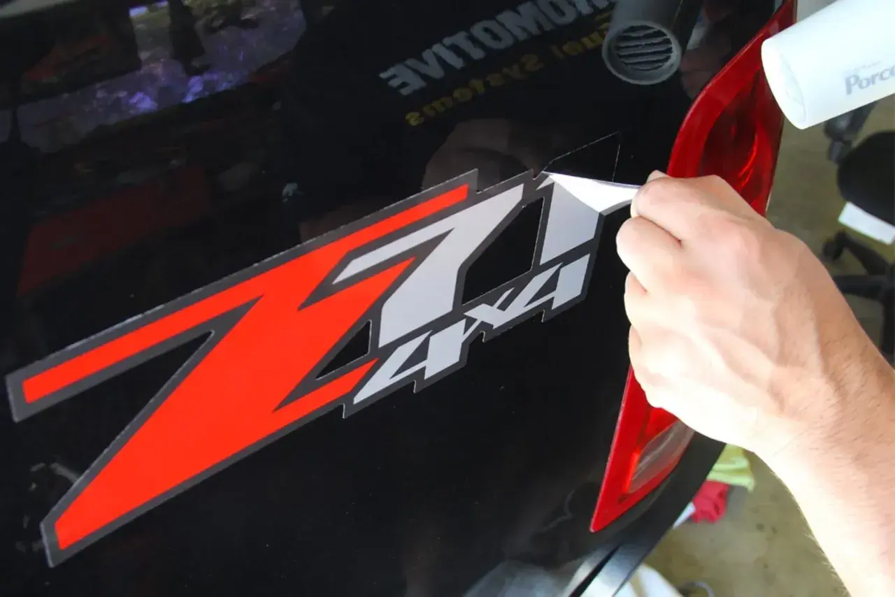 Mano che rimuove adesivo "Z7 4x4" da auto nera, usando un phon per ammorbidire il nastro adesivo vecchio.