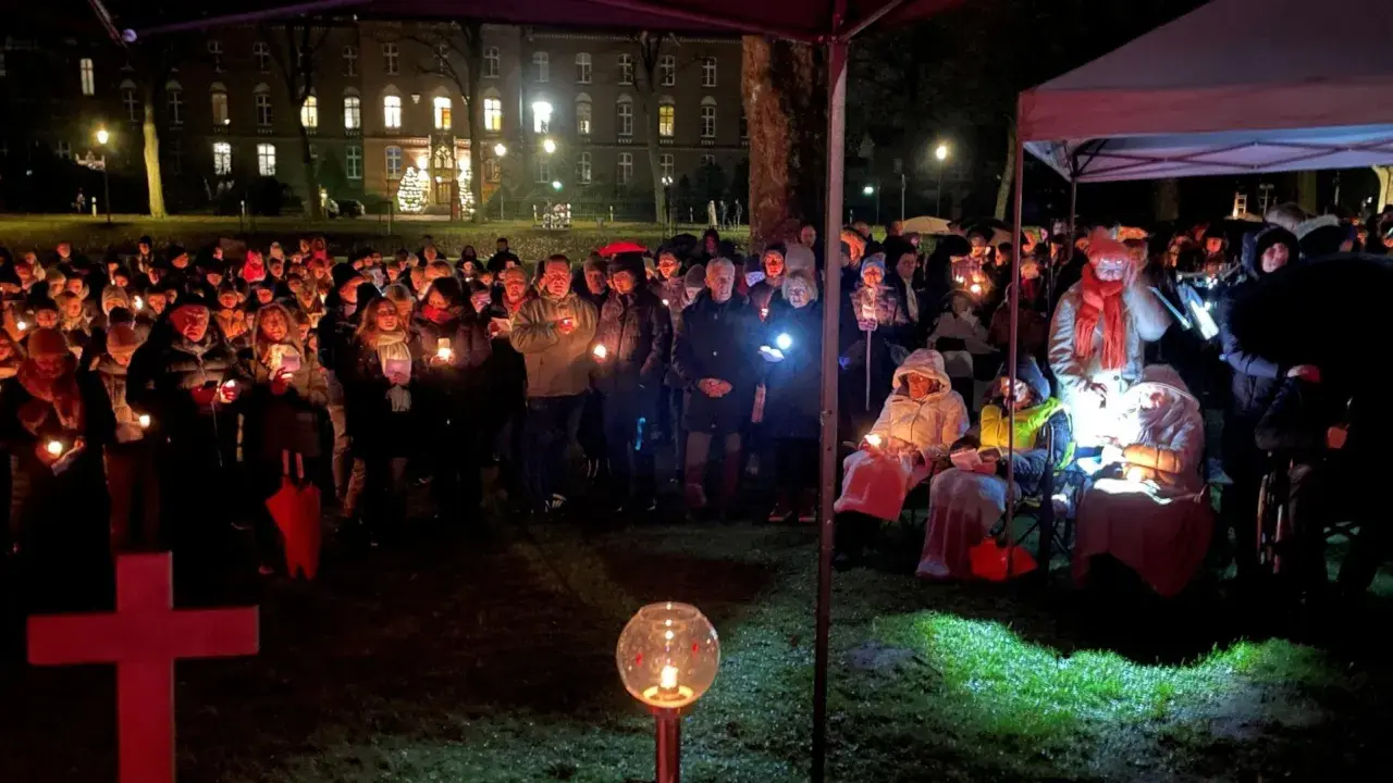 Menschen versammeln sich bei Kerzenlicht zu einem Gottesdienst am Heiligabend vor einem Gebäude. Ein rotes Kreuz steht im Vordergrund.