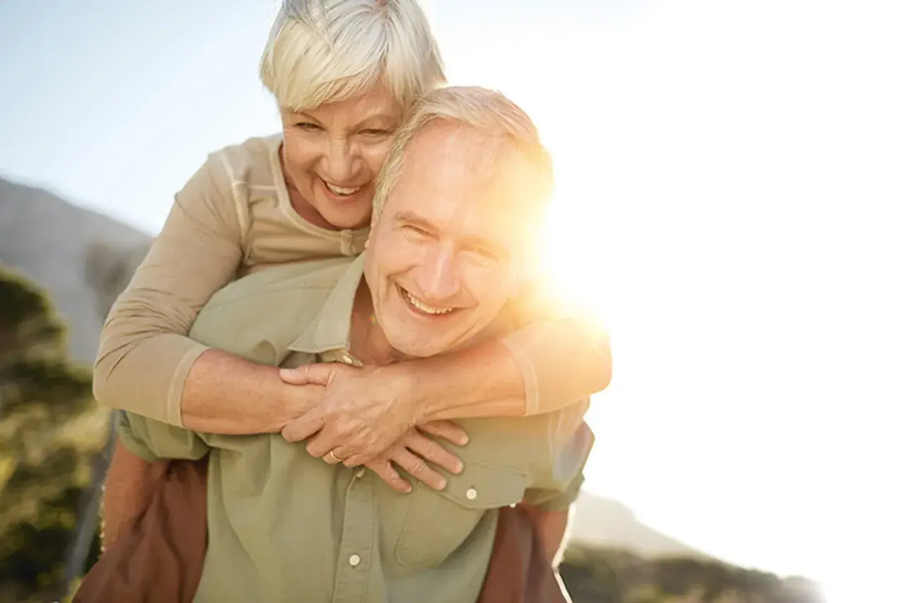 Ein Mann trägt eine Frau auf dem Rücken. Beide lachen und genießen die Sonne. Ein Bild von wahrer **Liebe im Alter**.