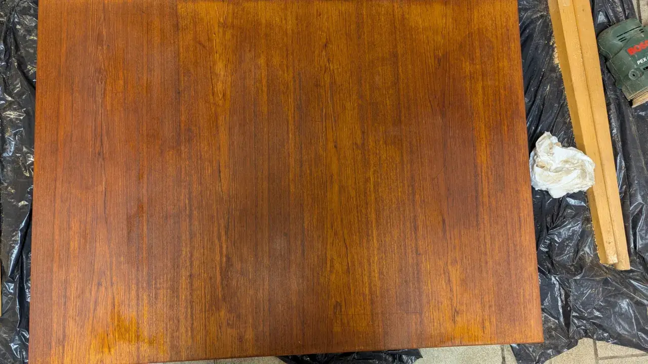 Förbereder för att slipa teak. En träyta ligger på svart plast, bredvid en slipmaskin och en trasa.
