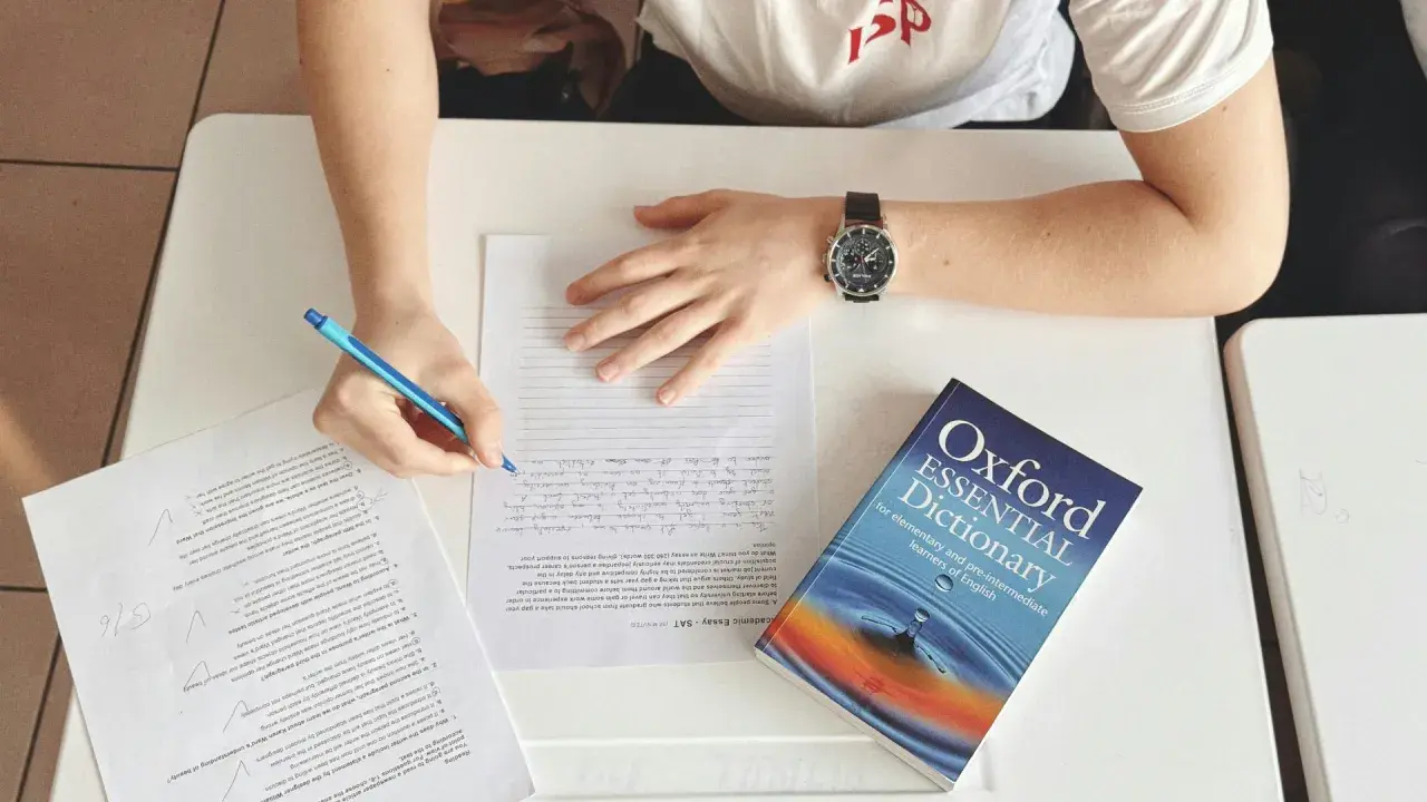 Studente che scrive, con un dizionario Oxford Essential Dictionary aperto. Cosa vuol dire AT? Forse "at" è un'abbreviazione usata in questo contesto di studio.