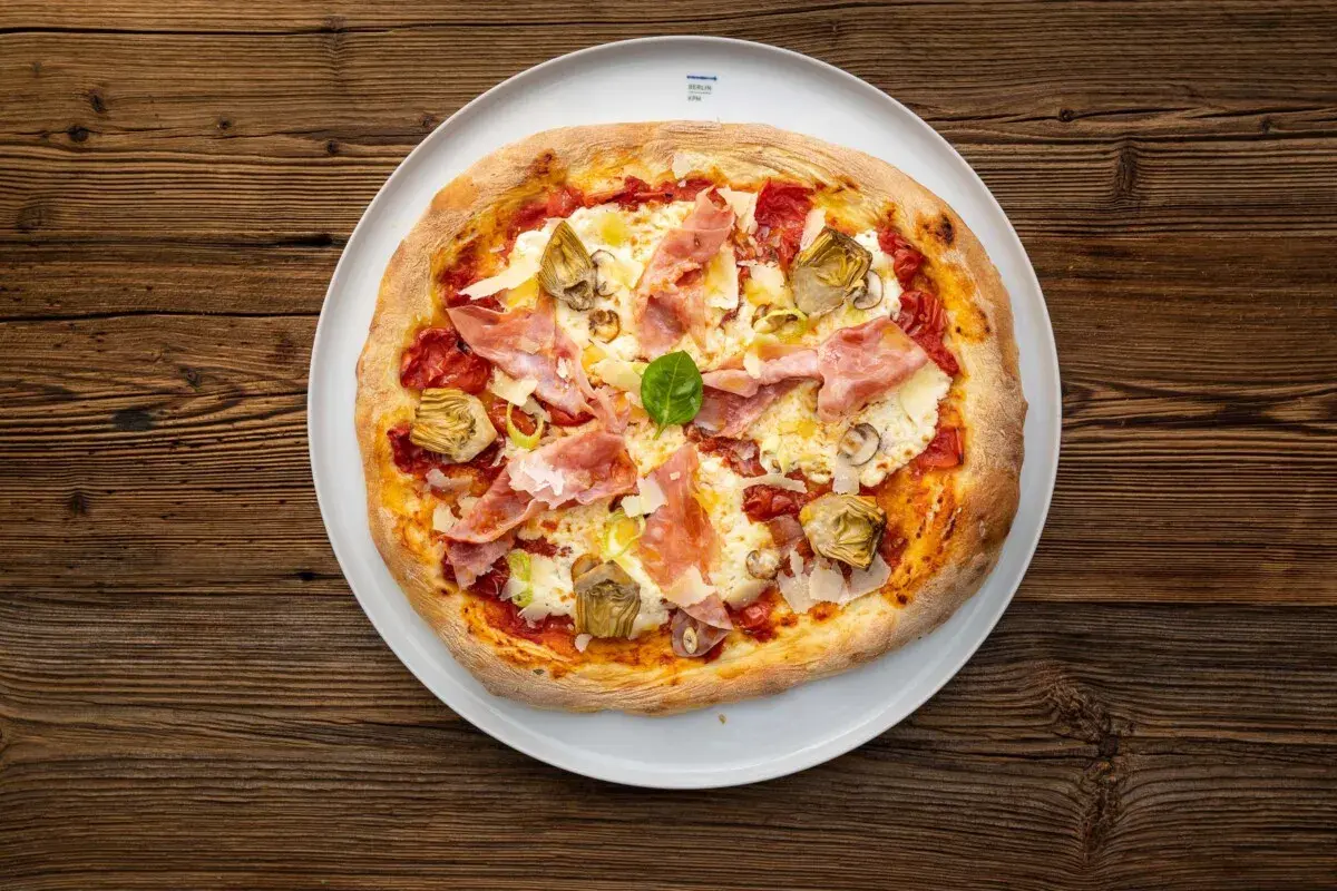 Eine köstliche Pizza mit Prosciutto Crudo, Artischocken und frischem Basilikum auf einem weißen Teller, serviert auf rustikalem Holz.