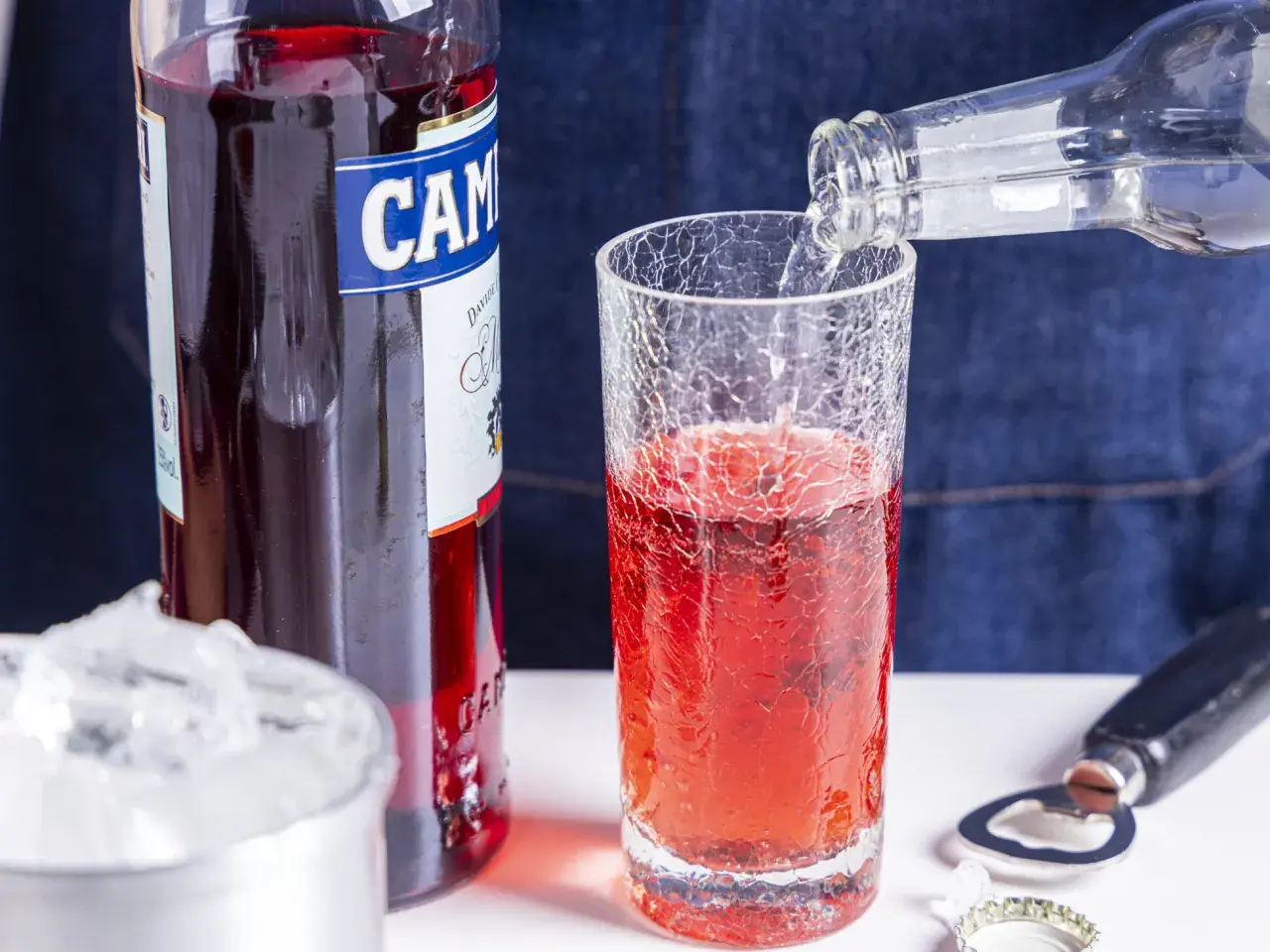 Campari Soda Rezept: Sprudelndes Wasser wird in ein Glas mit Campari gegossen. Eiswürfel und Flaschenöffner liegen bereit.