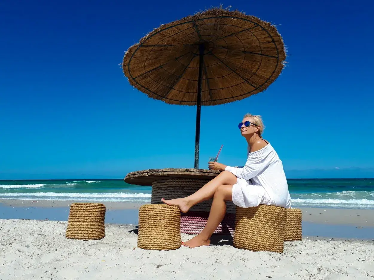 Kobieta w białej sukience siedzi pod parasolem na plaży na Djerbie. Uważać na słońce i upał.