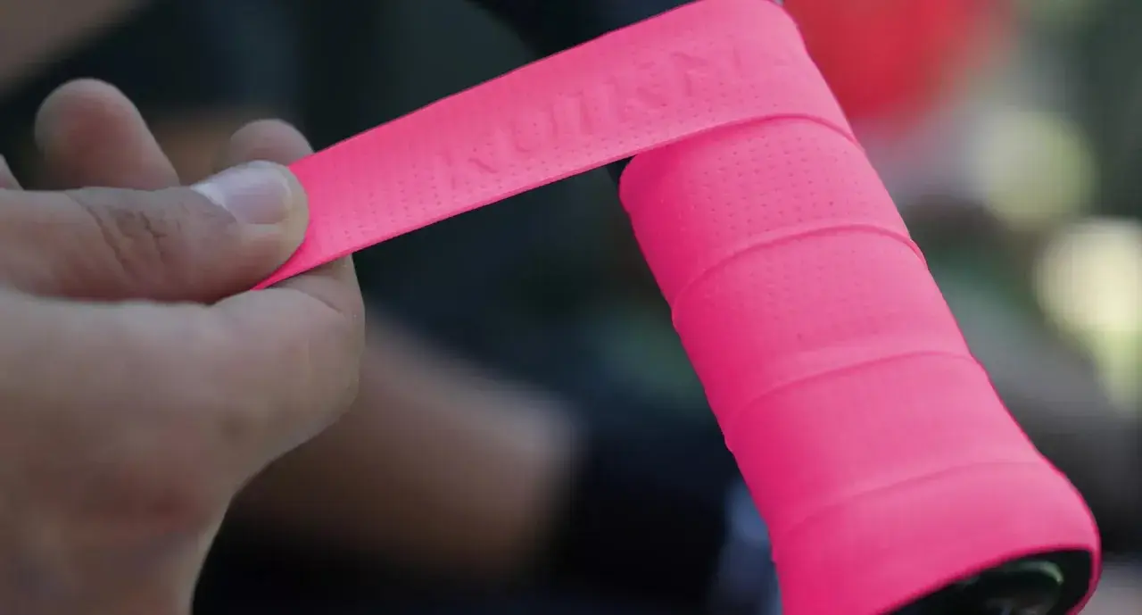 Mano applica nastro rosa su manubrio bici, mostrando la differenza tra grip e overgrip.
