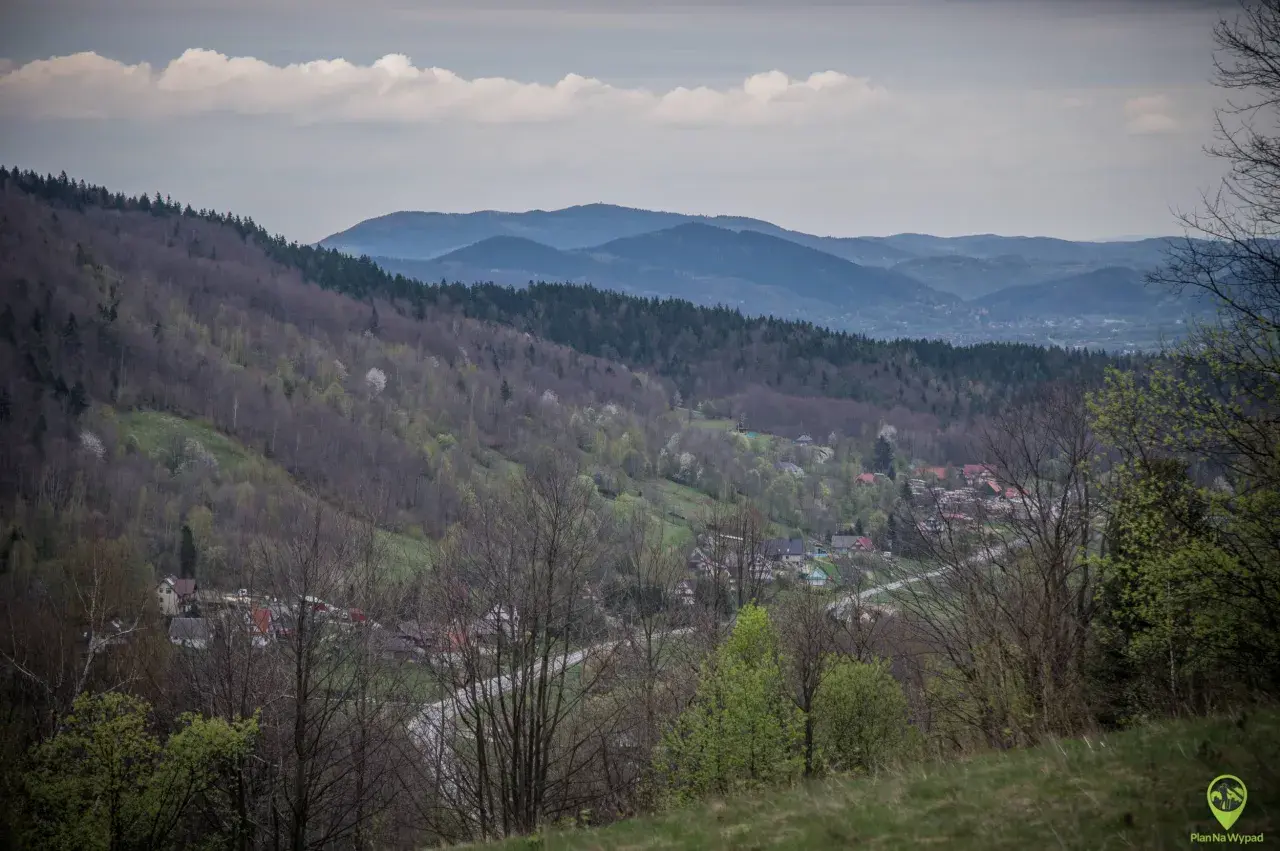 Widok na dolinę z zabudowaniami, lasami i pasmem górskim w tle. Widać tu ćwilin z Jurkowa, wiosenną zieleń i chmury na niebie.