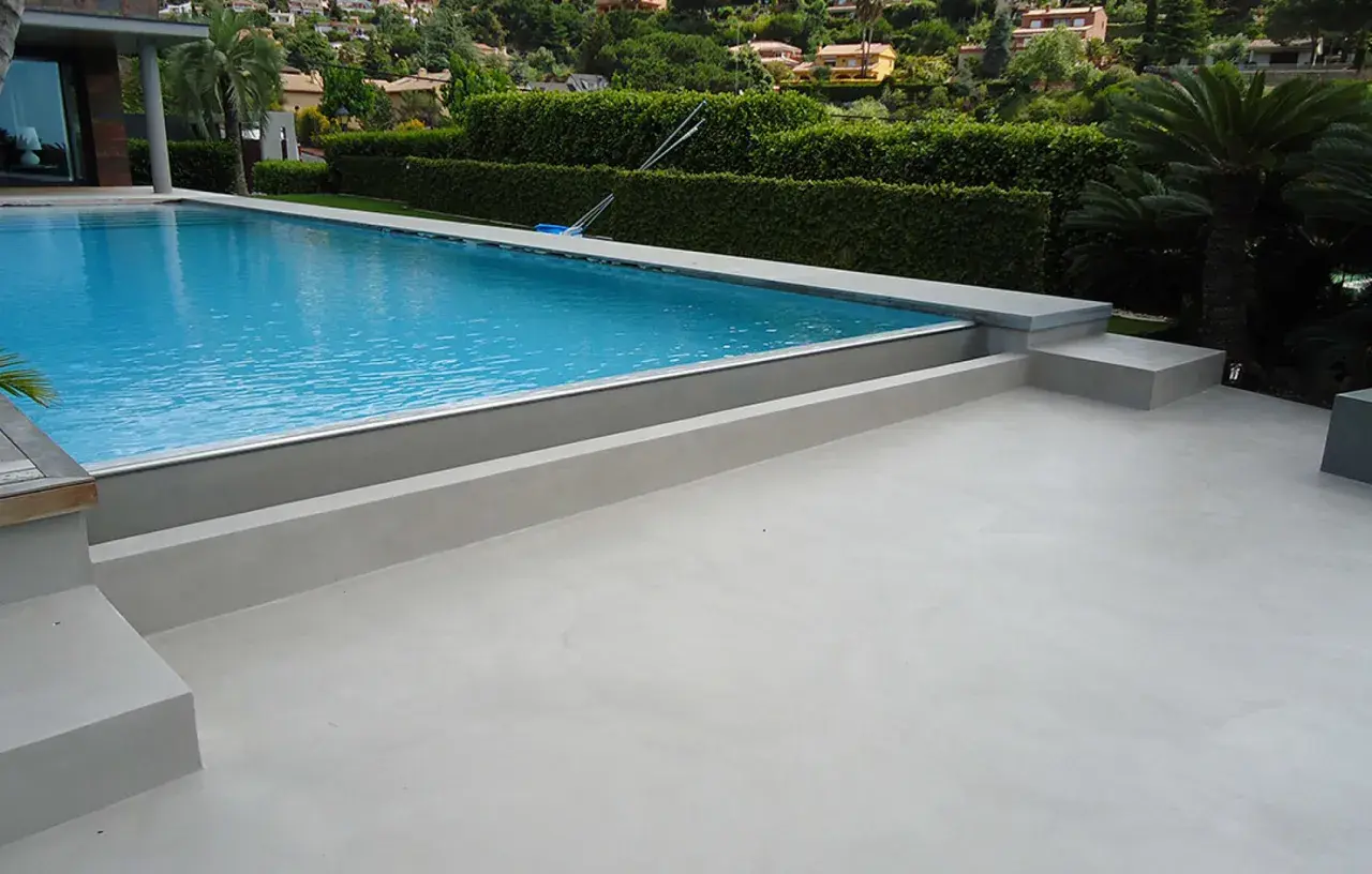 Modern villa with a bright blue pool and surrounding terraces finished with elegant piscinas de microcemento. Lush green hedges and palm trees frame the scene.