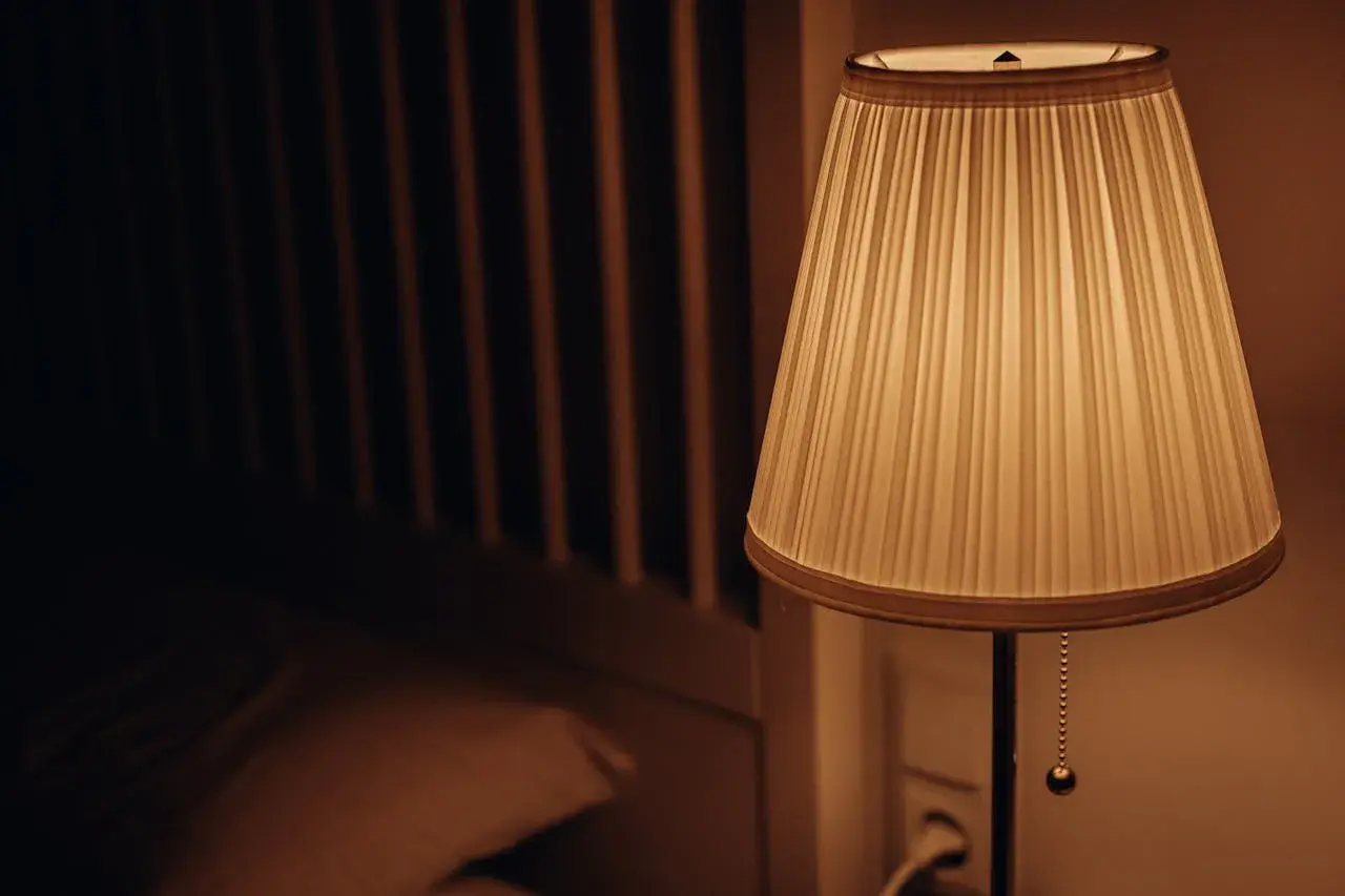 Lámpara con pantalla cilindrica de tela plisada, iluminando suavemente una habitación oscura con una cama al fondo.