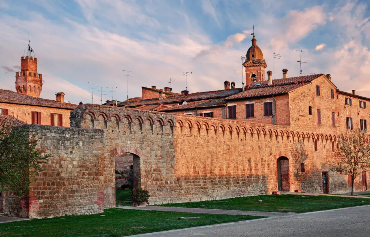 Antica cinta muraria in pietra delle Crete Senesi, con torri medievali e cielo al tramonto.