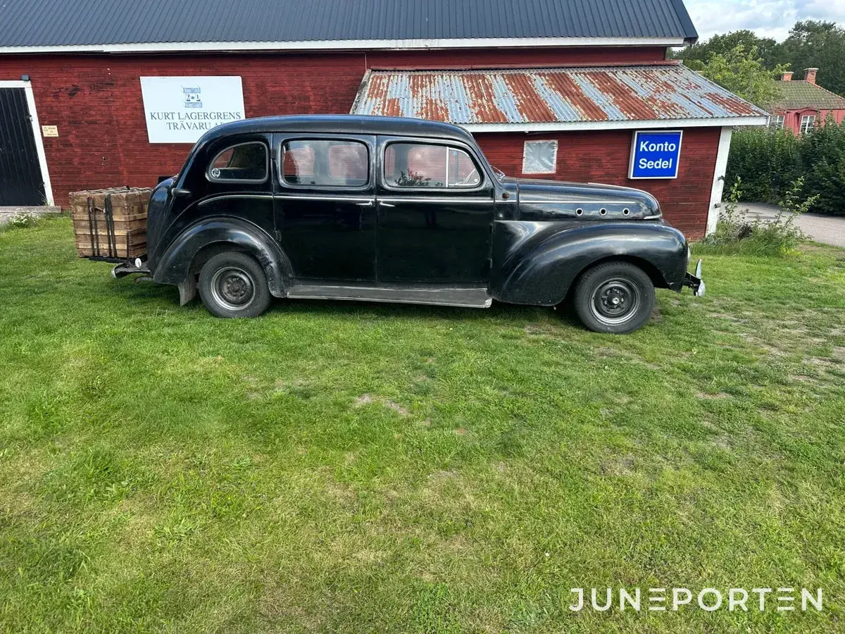 Svart Volvo PV 831 parkerad på gräs framför en röd byggnad med rostigt plåttak. En träkorg sitter på bakre stötfångaren.