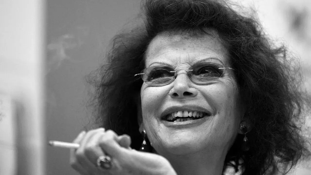 Uśmiechnięta Claudia Cardinale, ikona kina, z papierosem w dłoni. Jej spojrzenie pełne jest życia, jak w jej najlepszych filmach.