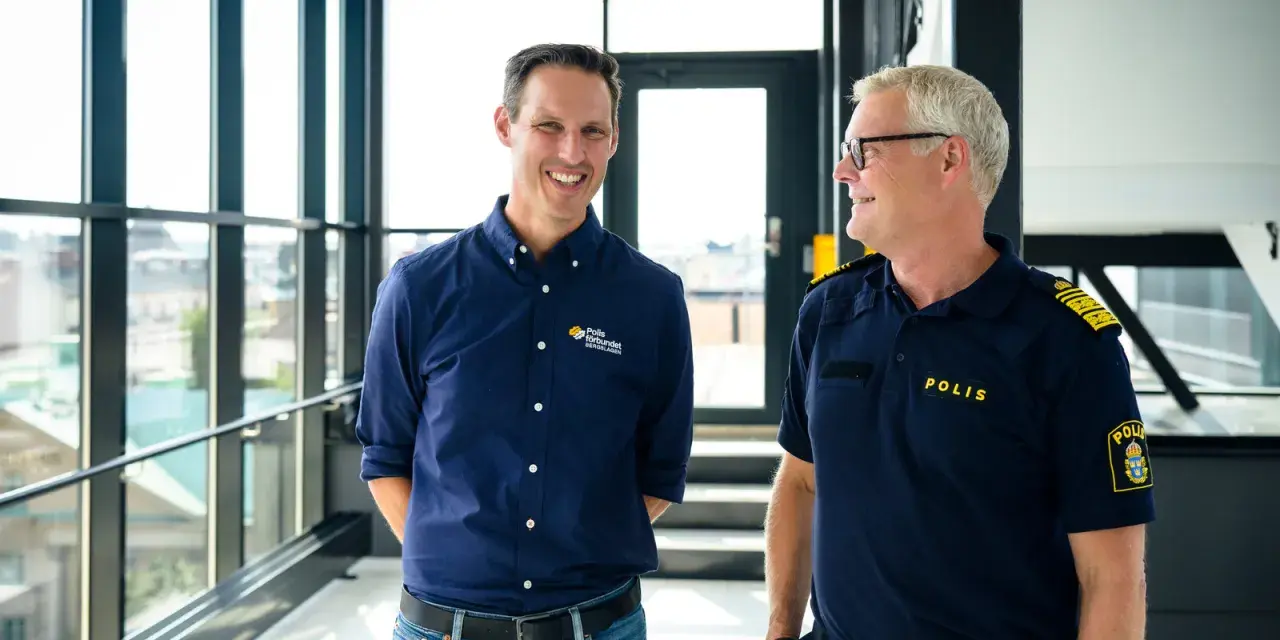 Två män ler mot varandra. Den ena bär en mörkblå skjorta med texten "Polis förbundet Bergslagen", den andra en polisuniform med texten "POLIS".