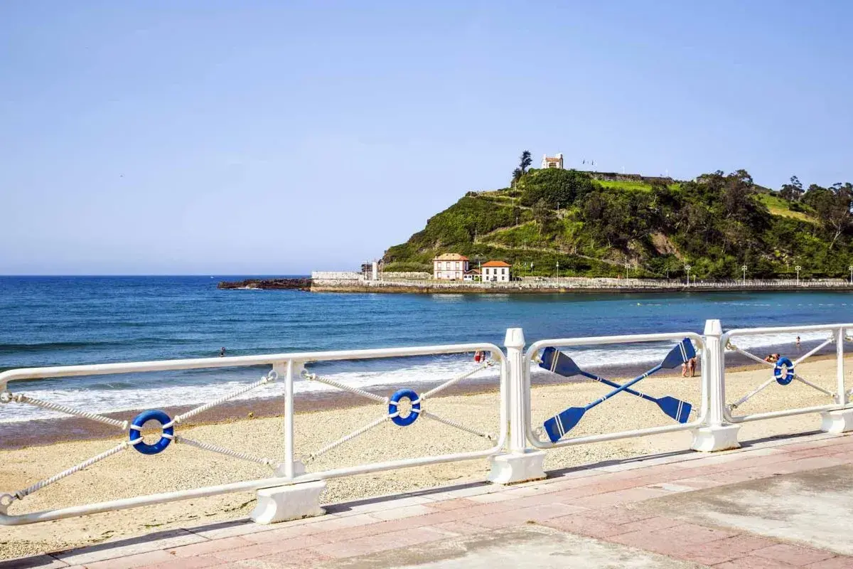 La **playa de Santa Marina** con su arena dorada, el mar azul y una colina verde coronada por un edificio blanco.