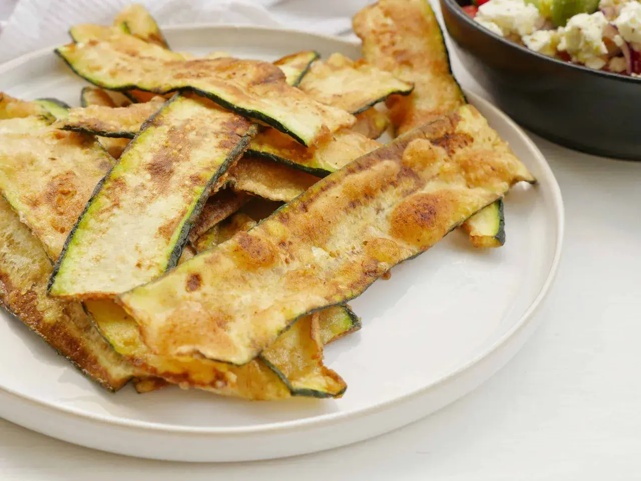 Krispiga, gyllene skivor av friterad zucchini ligger staplade på en vit tallrik, redo att avnjutas som ett lätt tilltugg.