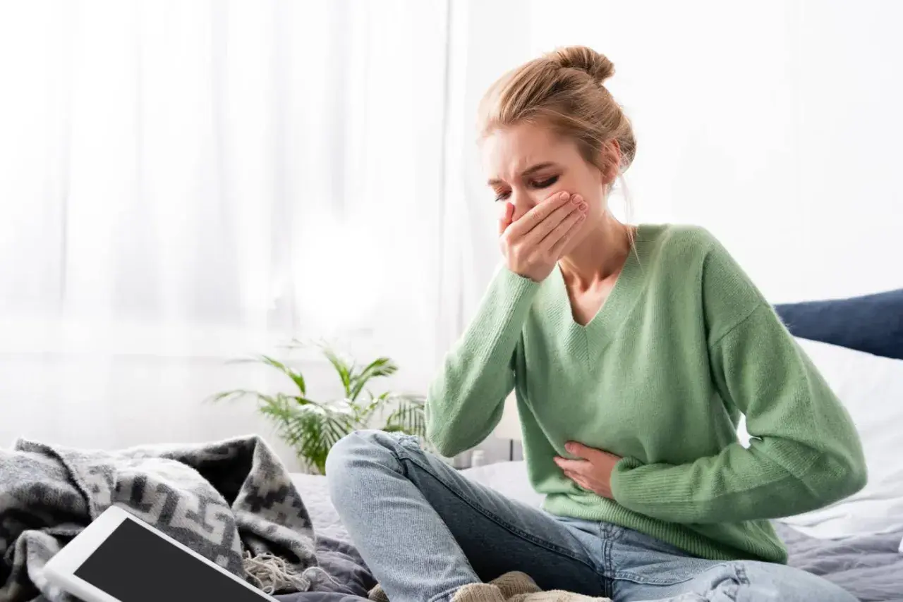 Mujer rubia con moño sentada en la cama, con la mano en la boca y otra en el abdomen, sufriendo nauseas antes de la regla.