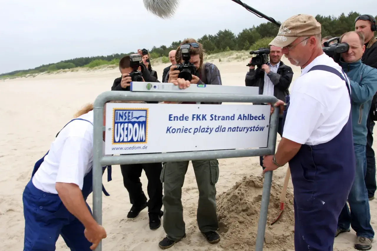 Na plaży nudystów stogi piasku i tablica informacyjna. Ludzie z aparatami dokumentują wydarzenie.