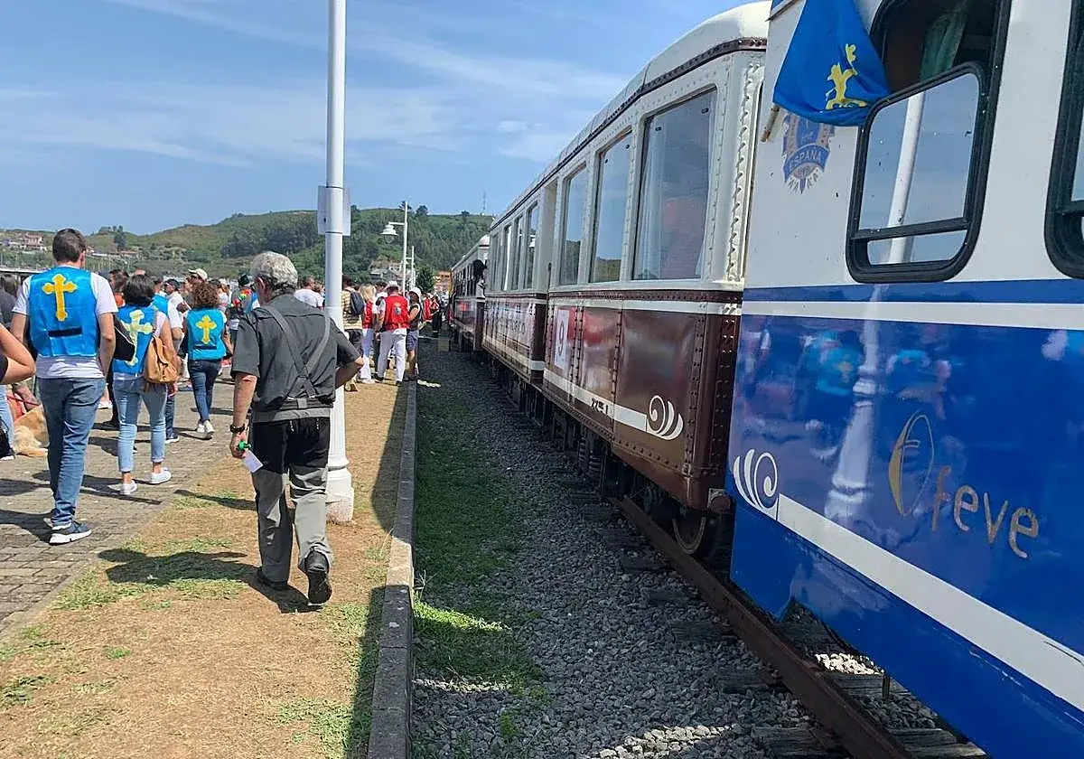 Gente con chalecos amarillos espera el tren descenso del Sella. El tren tiene vagones marrones y azules con detalles dorados.