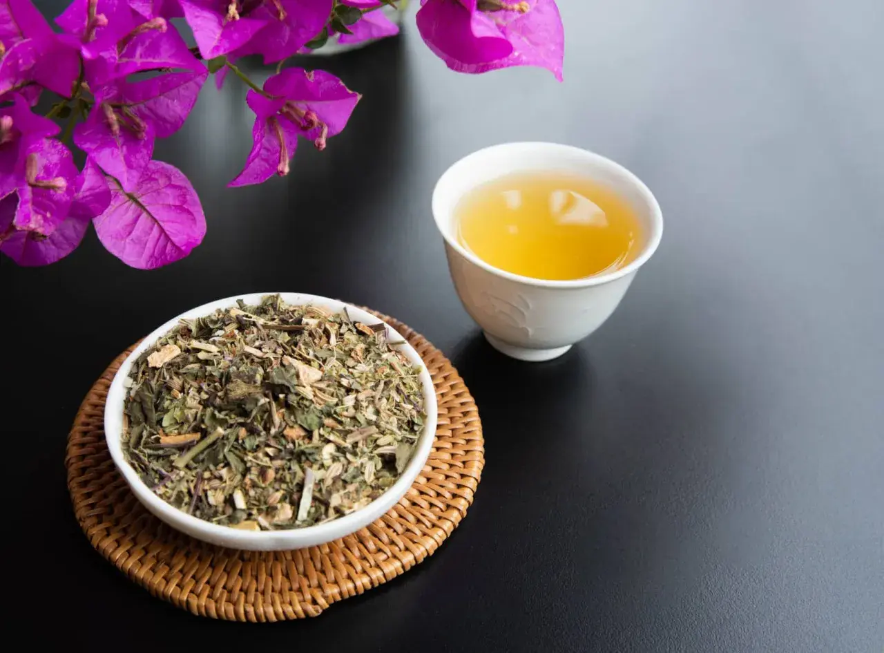 Una tazza di tisana per digerire accanto a una ciotola di erbe secche, con fiori viola sullo sfondo.