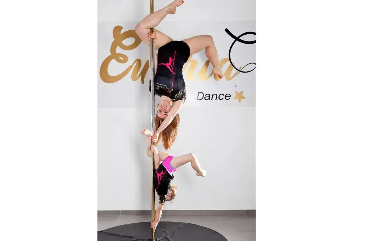Dwie dziewczyny ćwiczą pole dance. Młodsza wspiera starszą, która wisi głową w dół.