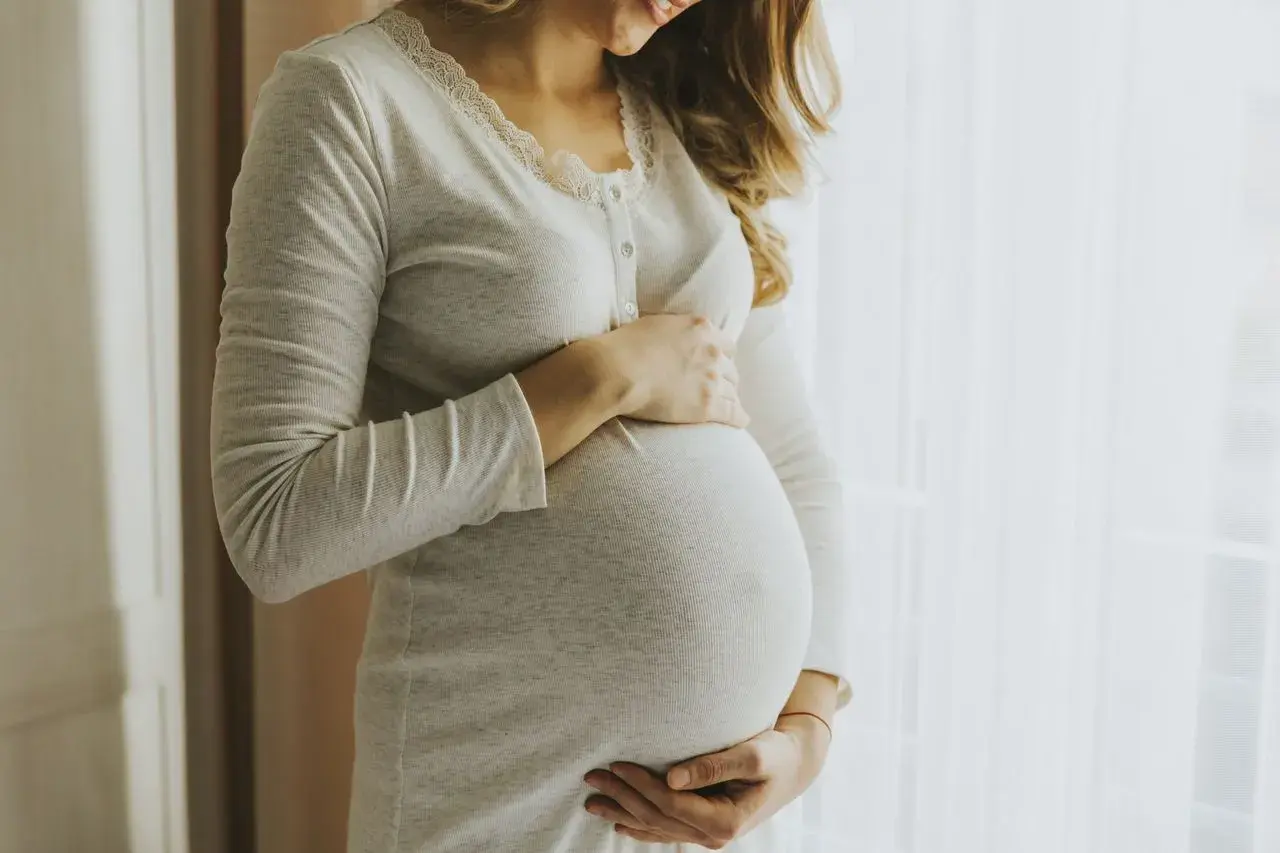 Kobieta w ciąży, z dłońmi na brzuchu, wygląda na szczęśliwą. Jej uśmiech sugeruje, że jest pełna życia, jak nimfomanka.