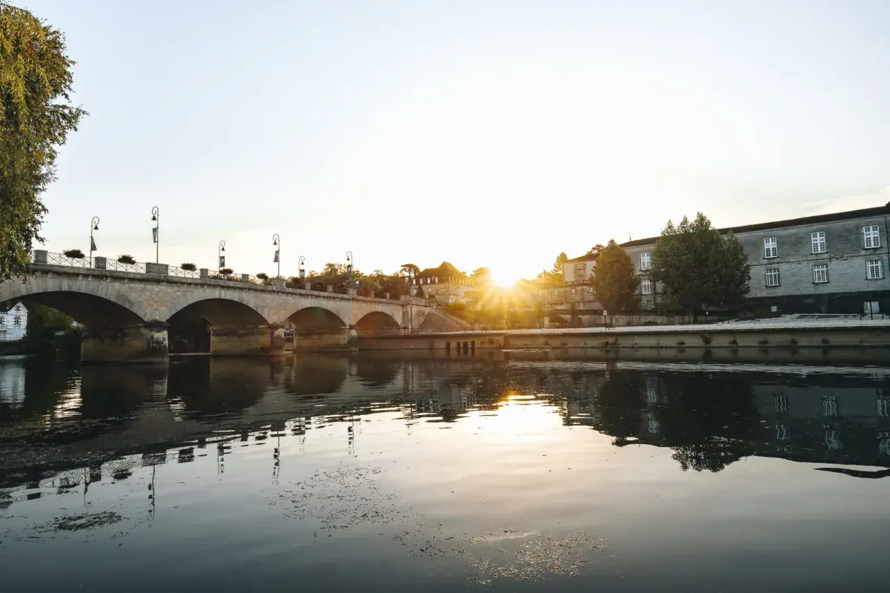 Un pont de pierre traverse une rivière calme, reflétant le soleil couchant sur les bâtiments historiques. Une belle vue pour savoir quoi faire à Cognac.