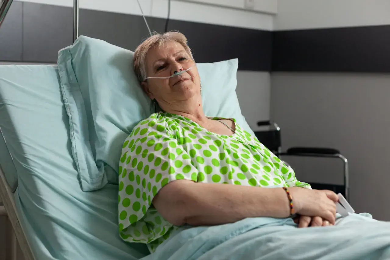 Starsza kobieta w szpitalnym łóżku, z tlenem przy nosie, w zielono-białej bluzce w groszki. Czeka na leki, być może Duracef.