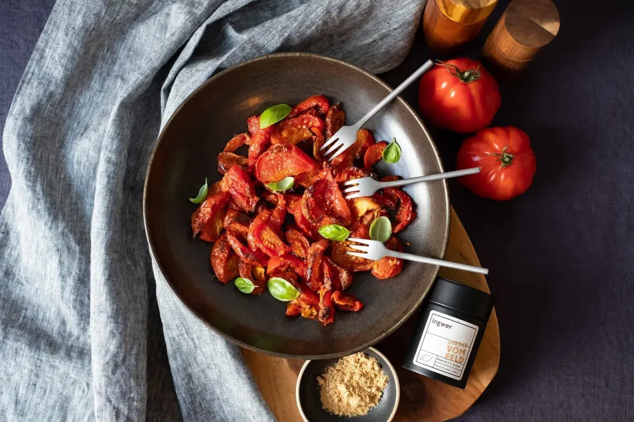 Schüssel mit karamellisierten Tomaten, garniert mit Basilikum. Daneben frische Tomaten, Ingwer-Gewürz und ein kleiner Napf mit Ingwerpulver.