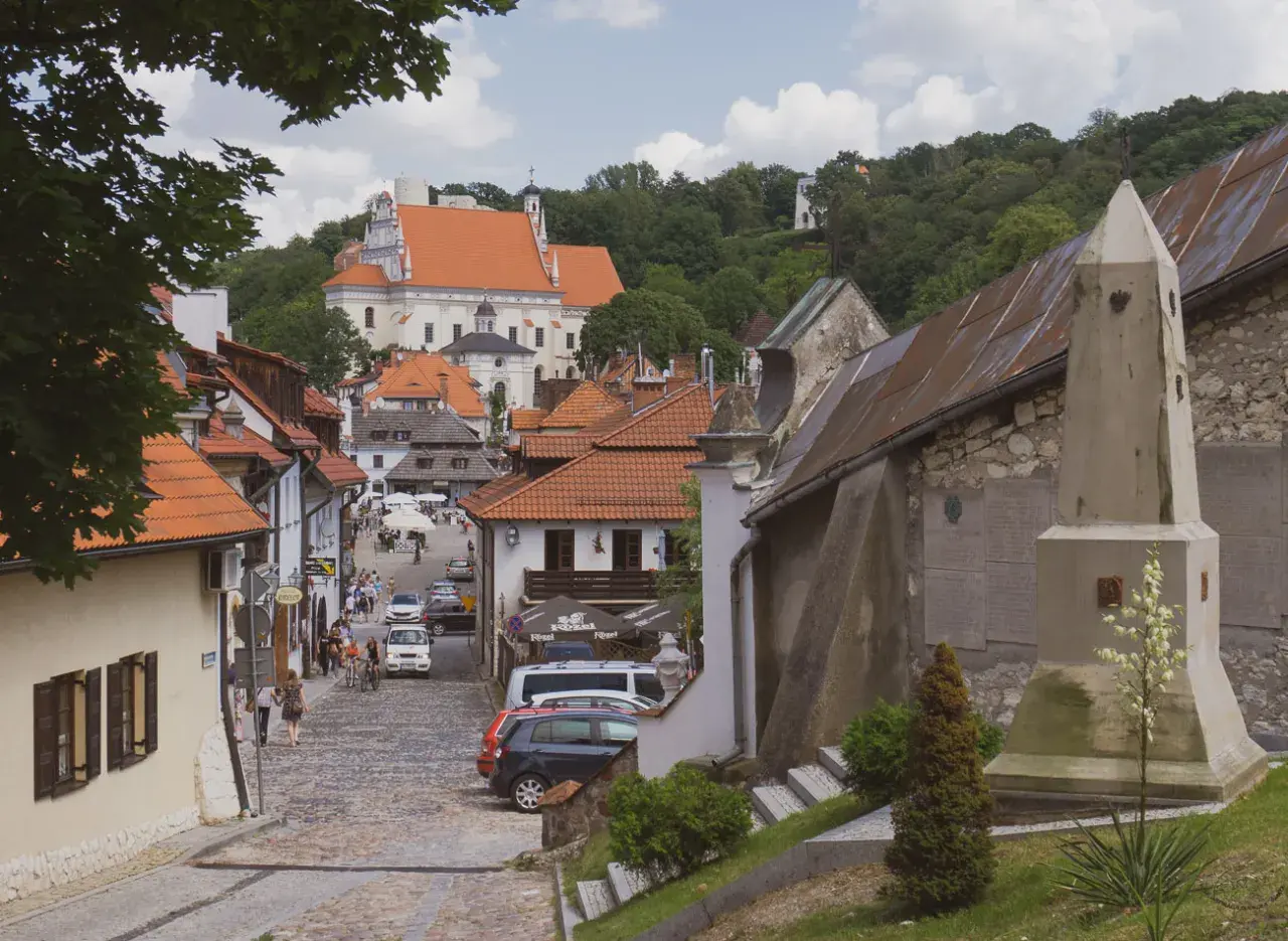 Urokliwa ulica w Kazimierzu Dolnym z kościołem na wzgórzu. Kamienne bruki, kamienice z czerwonymi dachami i zielone drzewa tworzą malowniczy krajobraz.